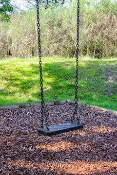An empty black swing hangs on long steel chains over a wood-chip surface in.. Stock Photos