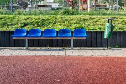 Empty blue seats line a waiting area by a playground in daylight, with a green Stock Photos
