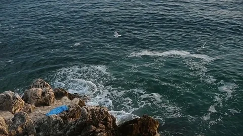 Empty blue towel on rocks by the sea and waves hitting rocks Stock Footage 76800579