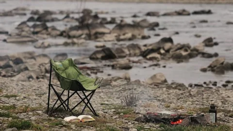 Empty camping chair by the river with an open book and campfire, wind turning Stock Footage 322202910