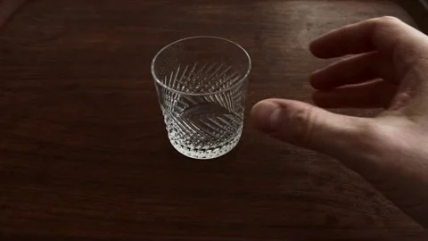 Empty crystal patterned glass is placed by hand on dark wooden table surface 動画素材 329621334
