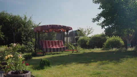 Empty Cushioned Swing Framed By Lush Garden And Open Lawn, Soft Sunlight On Vidéo 324618133