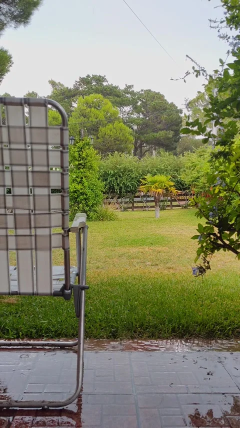 Empty folding chair sits on a wet garden, overlooking a lush green garden Stock Footage 308418966