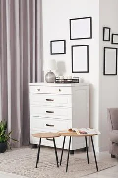 Empty frames hanging on white wall, chest of drawers and wooden tables indoor Stock Photos