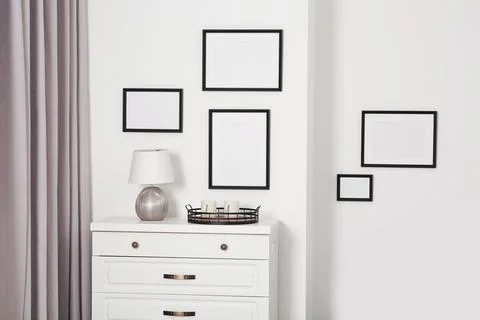 Empty frames hanging on white wall, wooden chest of drawers and lamp indoors Foto stock