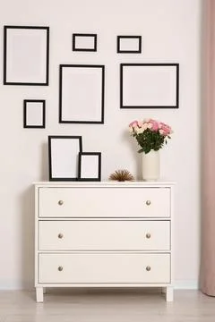 Empty frames hanging on white wall and chest of drawers with flowers in room Stock Photos