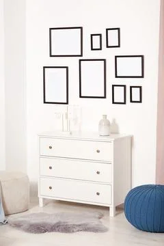 Empty frames hanging on white wall, chest of drawers and ottomans indoors Stock Photos