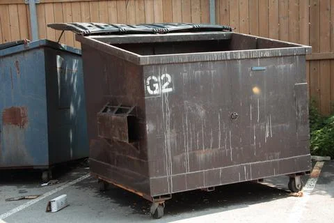 Empty garbage dumpster with lid open and another dumpster behind it, surrounded Stock Photos