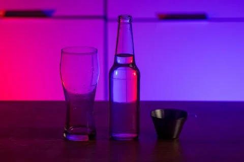 Empty glass bottle with clear liquid and metal measuring cup under neon lighting Stock Photos