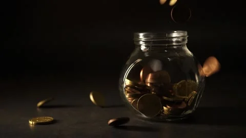 An empty jar on a dark table coins begin to fall inside and all over it Stock Footage 160557165