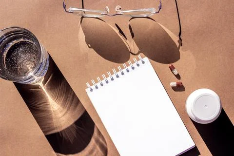 Empty notepad, pills, glass of water and eyeglasses on brown background. Crea Stock Photos