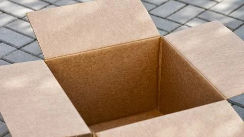 An empty open cardboard box sitting on a grey paved outdoor surface. Stock Photos