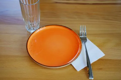 Empty orange plate with fork and glass on wooden table in bright setting Stock Photos