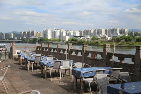 Empty outdoor dining tables beside river railing with residential skyline a.. Stock Photos