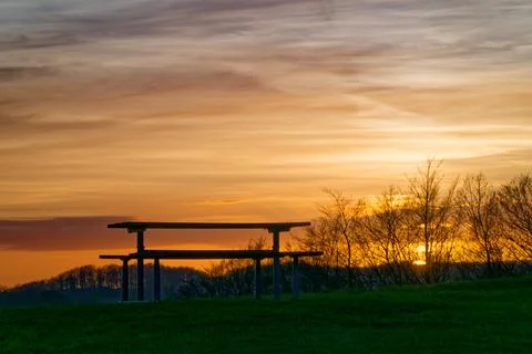 Empty picnic table on grassy hill during vibrant sunset with silhouetted tree Stock Photos