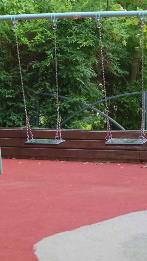 Empty playground swing set sways gently in the breeze, surrounded by lush green Vídeos de archivo 341439304