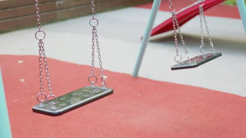 Empty playground swings gently sway in the breeze, with red rubber flooring and Видео 340901427