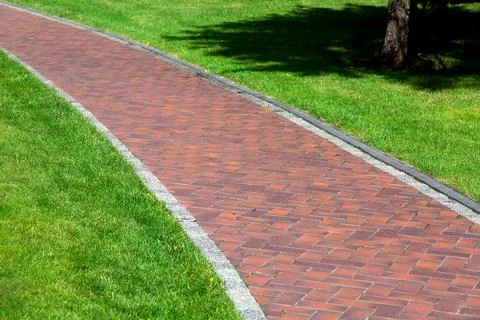 Empty red tile footpath in a park with a green lawn and a drainage system. Stock Photos