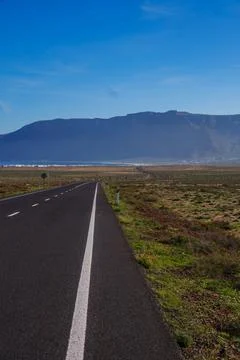 Empty road leads through vast green fields to dark mountains under br. Lanzarote Stock Photos