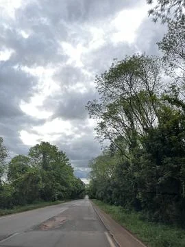 Empty road surrounded by lush green trees under a dramatic cloudy sky in Bo.. Stock Photos