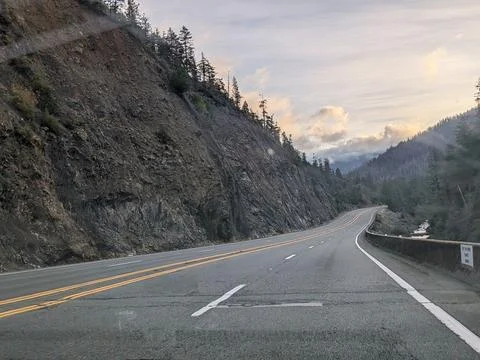 An empty rural highway winds between rocky mountains and a peaceful river Stock Photos