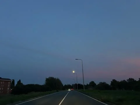 Empty rural road at dusk with glowing streetlights, distant car lights, and Stock Photos
