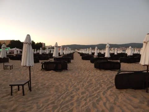 Empty sandy beach with folded white umbrellas and brown sunbeds at sunset Stock Photos