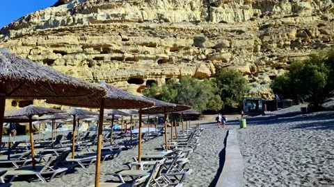 Empty Sun Loungers Under Straw Parasols Below Cliffside Caves in Crete Stock Footage 337538244