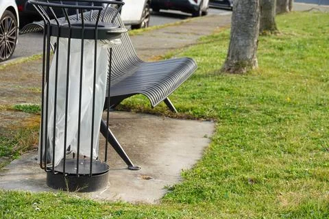 Empty trash can next to public bench on city sidewalk surrounded by grass Stock Photos