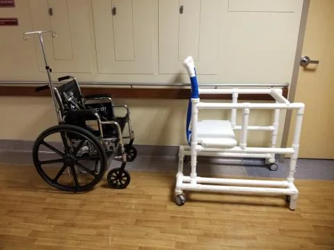 Empty wheelchair and bedside commode lined up in hospital hallway Foto stock