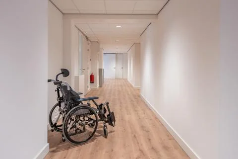 An empty wheelchair stands in an empty corridor of a care home with white wal Stock Photos