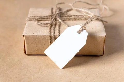 Empty white gift tag mock up on craft paper box on natural beige background. Stock Photos