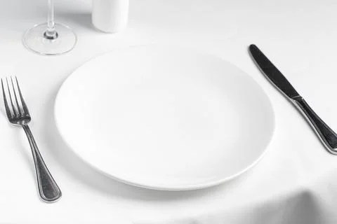 Empty white plate with fork and knife on white tablecloth in restaurant. Mo.. Stock Photos