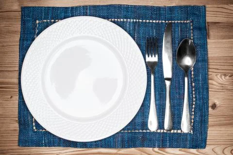 Empty white plate, fork, knife and spoon  on blue placemat over a wooden tabl Stock Photos
