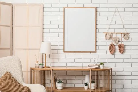 Empty wooden frame on a white brick wall with a console table, lamp, and deco Stock Photos