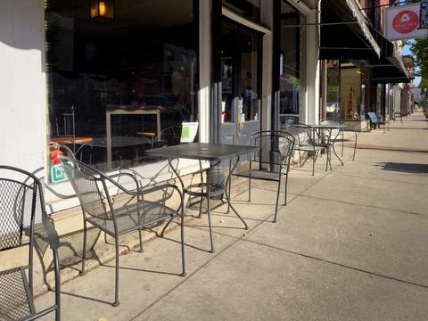 Empty wrought-iron mesh patio tables and chairs along a sunlit sidewalk Stock Photos