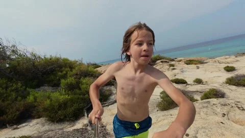 Energetic shirtless boy running fast on a rocky coastal trail with sea in the Video stock 313842616