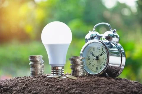 Energy saving light bulb and alarm clock on stacks of coins on nature backgro Stock Photos