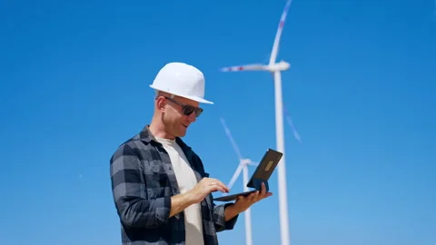 Engineer checks data on laptop at wind farm during sunny day. A person wearing a Stock Footage 342737212
