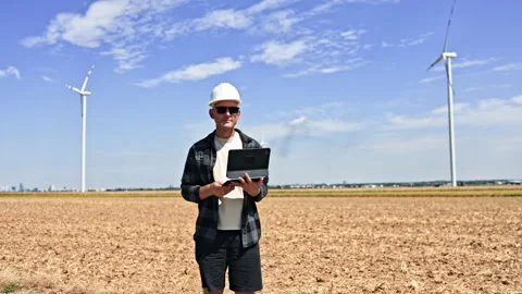 Engineer checks a tablet in a harvested field beside wind turbines during Stock Footage 348694764
