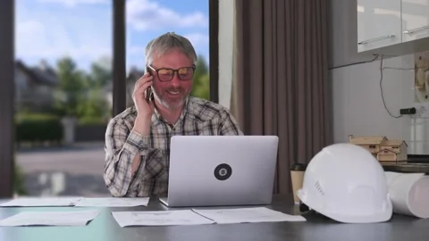 Engineer discussing project details by phone at kitchen table with laptop and Stock Footage 334870972