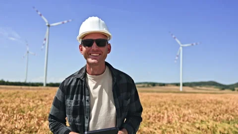 Engineer in a field checks a tablet beside wind turbines under a clear sky Stock Footage 348693529