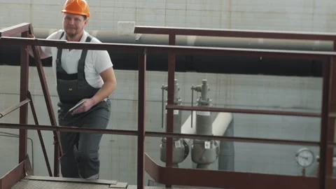 An engineer in a hard hat and uniform climbs a metal staircase at a factory. Stock Footage 306818763
