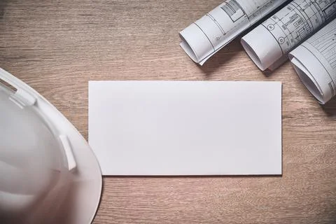 Engineer hardhat, rolled drawings and blank white envelope at wooden table Stock Photos