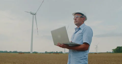Engineer in hardhat uses laptop near wind turbines environmental Stock Footage 216698677
