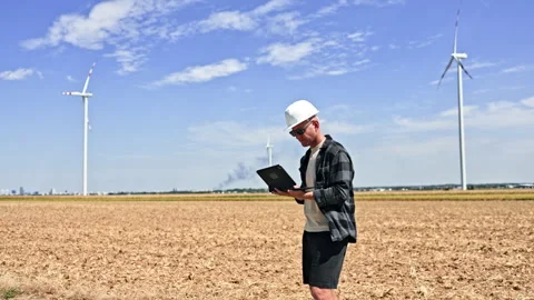 Engineer holding a tablet in a plowed field beside wind turbines during the day Stock Footage 348695049
