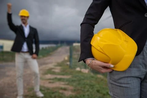 An engineer holds a hardhat in one hand while her colleague behind celebrat.. Stock Photos