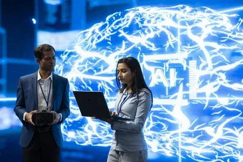 Engineer holds VR headset while colleague uses laptop in front of AI brain Stock Photos