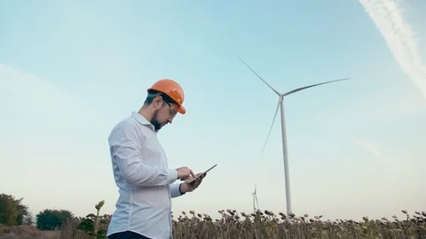Engineer in orange hard hat at windmill power plant with tablet in hand on sky Stock Footage 99582507
