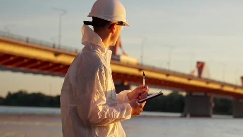 Engineer in protective gear standing near a large bridge at sunset, taking Stock Footage 323337681